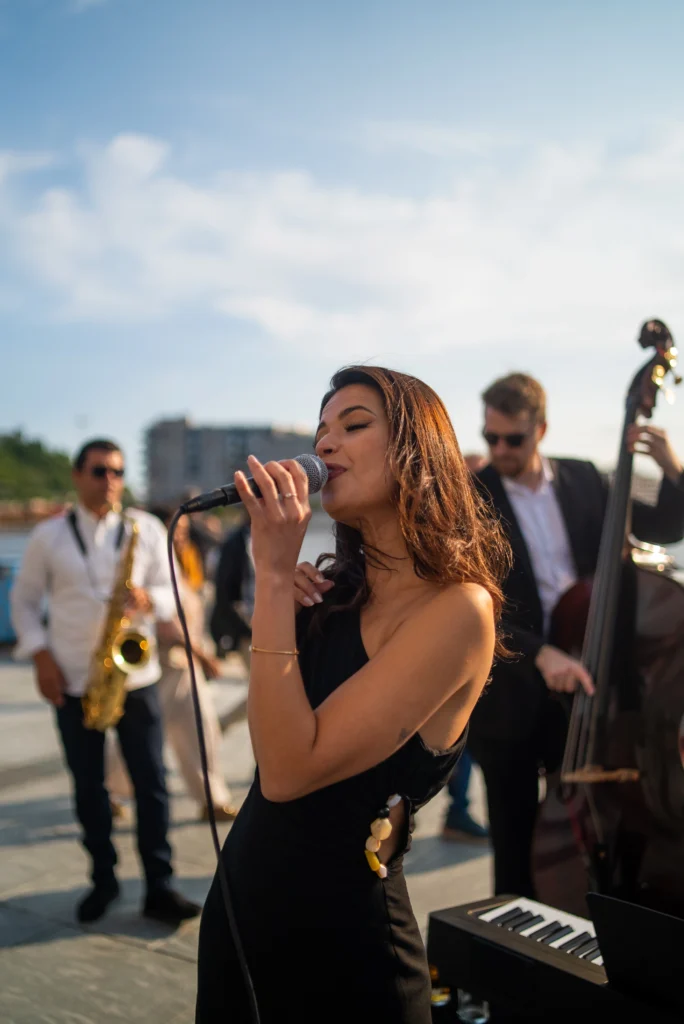 A female jazz singer performing outdoors with a jazz ensemble, featuring a saxophonist and double bass player in the background.