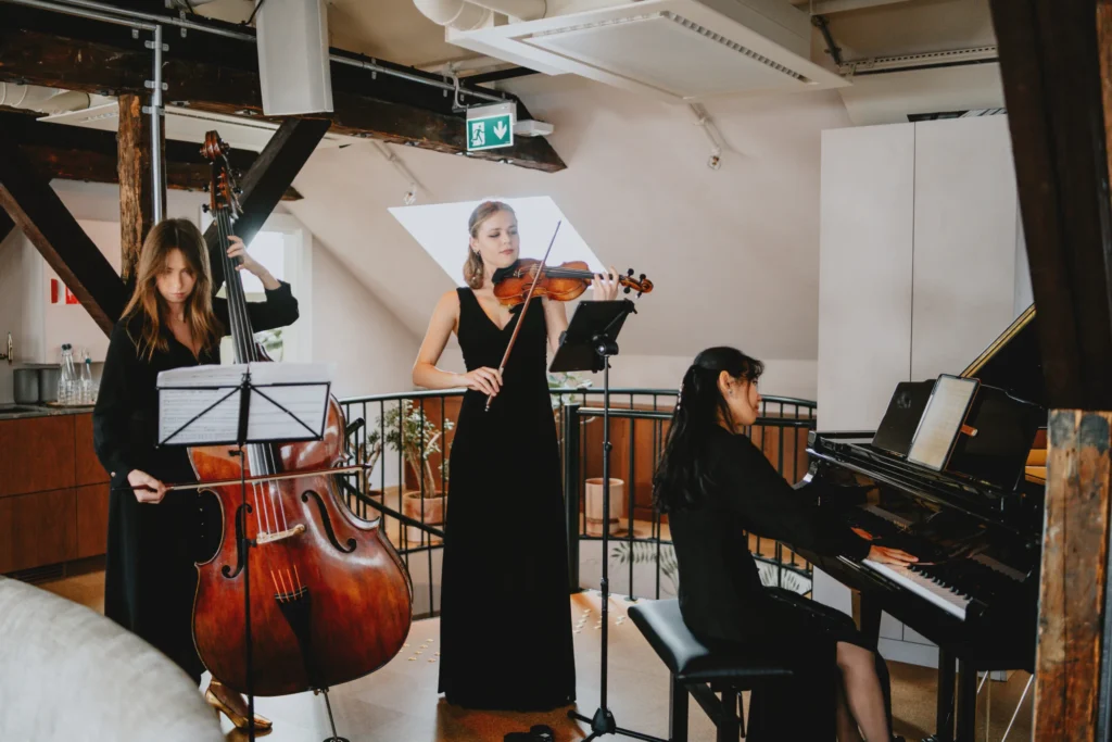 En trio jenter i svarte kjoler som fremfører sofistikert musikk ved et minnesmerke, med en på piano, en på cello og en på fiolin.